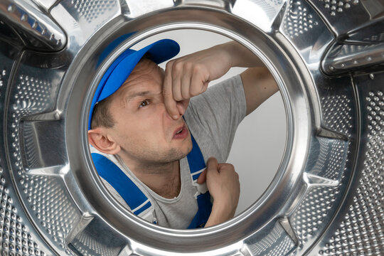Male repairman in uniform looks into the drum of a washing machine and covers his nose from an unpleasant smell, photo from the inside