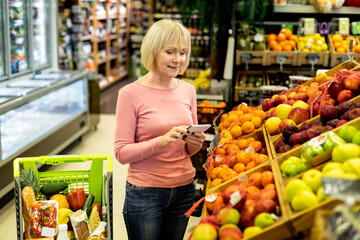 Attractive elderly woman choosing fruits at supermarket, holding notepad