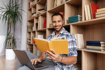 Happy arab man reading book near laptop computer, learning online while sitting at home