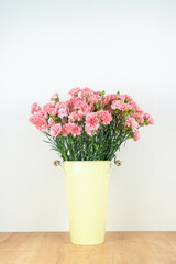 Close-up of a lot of pink carnations in a tin vase with handles on a wooden table on a neutral background. Vertical photo