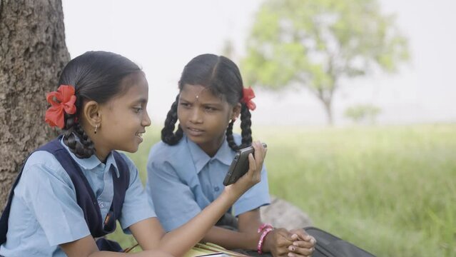handheld shot of village girl kids watching online class on mobile phone by sharing near farmland - concept of e-learning, education and rural lifestyle