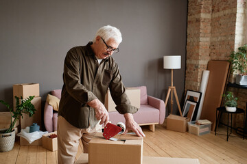 Horizontal medium shot of mature man preparing for moving to new house packing stuff into boxes and taping them