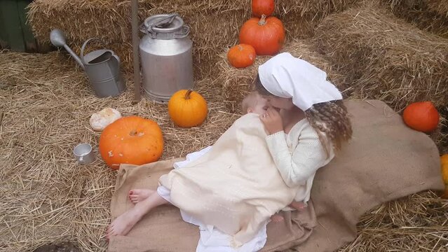 A young mother is breastfeeding her baby, sitting on a linen cloth in stacks of straw against the backdrop of pumpkins and rural accessories