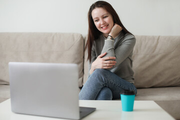 Young woman using video chat on laptop sitting on cozy sofa at home, watching video, having fun