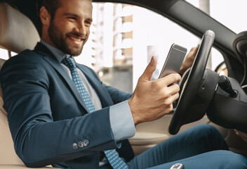 Adorable Caucasian businessman in suit looking to the screen of smartphone in an automobile