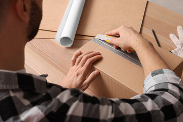 Man using utility knife to open parcel at wooden table, closeup - obrazy, fototapety, plakaty