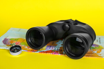 Modern binoculars, compass and map on yellow background, closeup