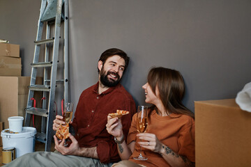 Horizontal shot of young man and woman in love sitting together on floor eating pizza and drinking...