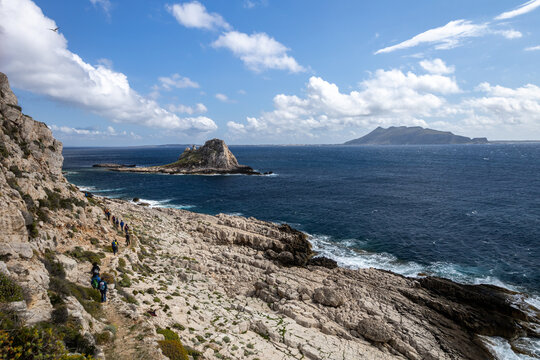 hikers walk toward the Faraglione (stack) on the island of Levanzo with Favignana in the background. (Egadi) Aegadian Islands, Trapani, Sicily, Italy