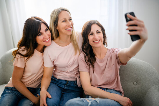 Three cheerful happy woman using mobile smart phone at home together sitting on sofa take a photo