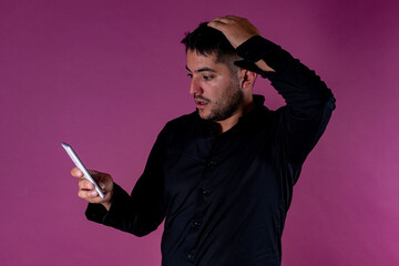 A man with a black shirt, posing on a pink background looking at the cell phone with a gesture of doubt and terror. Emotions, joy and fun of men.