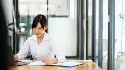 Young beautiful smile Asian businesswoman using her laptop while sitting in a chair at her working place.