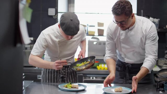 Sous chef serving cooked vegetables with fish on plates while helping head chef in restaurant kitchen
