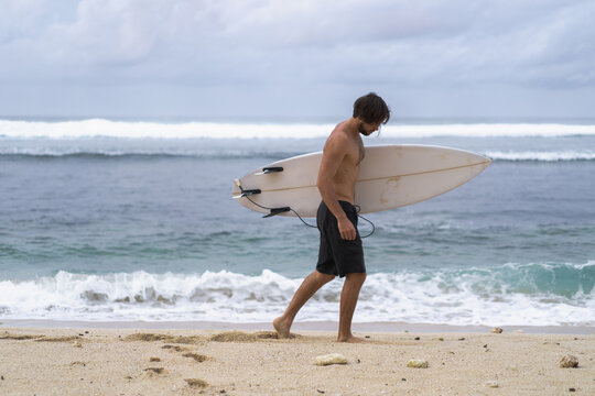 A young handsome man surfer on the ocean shore is doing a warm-up before surfing. exercises before sports, stretching before surfing.