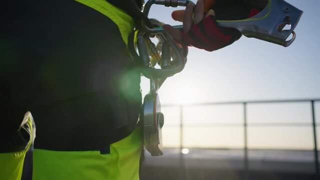 Close-up of an industrial climber fastening a carabiner with a jumar to a safety belt in front of the sun
