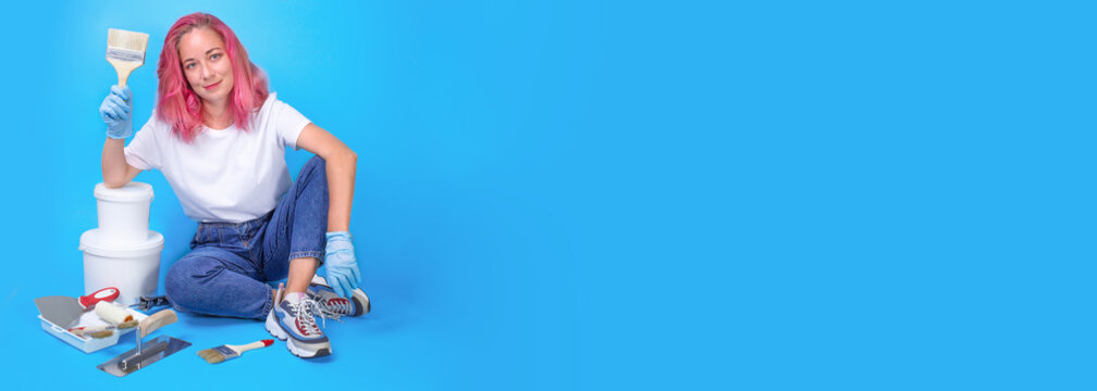 Banner. Smiling young woman with paint brush is going to do repairs on blue background. Instruments accessories for renovation apartment room. Repair home concept, construction and profession