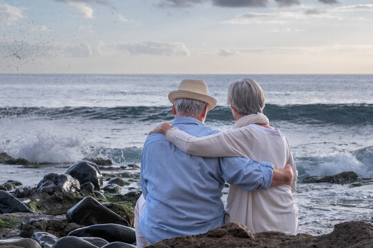 Rear View Of Senior Caucasian Couple Sitting On The Pebble Beach Hugging Looking At Horizon Over Water. Elderly Relaxed Couple Enjoying Nature And Freedom In Vacation Or Retirement