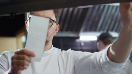 Professional male chef in glasses and uniform reading order slips while working in restaurant kitchen