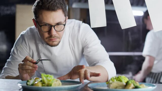 Professional male chef in glasses and uniform serving cooked dishes with tweezers on plates while working on food order in restaurant kitchen