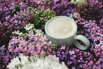 Fototapeta na wymiar Stylish composition of lilac flowers and warm cup of coffee. Colorful lilac branches and coffee with creamy foam, good morning concept. Spring morning in countryside still life