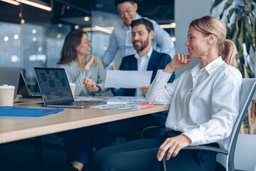 Smiling confident female boss sitting on meeting and looking at side with her colleagues at background in office. Business concept