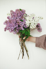 Stylish woman holding beautiful lilac flowers against white wall. Female in linen dress with lilac flowers, cropped view. Authentic moody moment. Rustic wedding