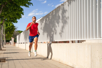 Young man running along a path between trees and a white fence. On a blue sky day. He is wearing a...
