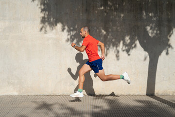 Young athletic Caucasian man running beside concrete wall. Shadow is cast on the wall. Wearing red T-shirt and blue shorts. Spain, Alicante.