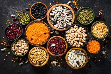 Legumes, lentils, chikpea and beans assortment in different bowls on black stone table. Top view.