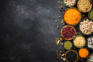 Legumes, lentils, chikpea and beans assortment in different bowls on black stone table. Top view.