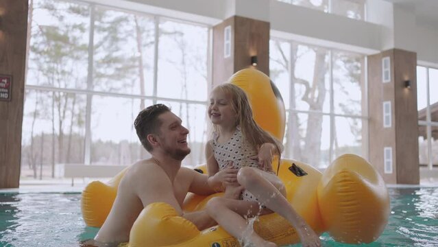 joyful little girl is laughing in swimming pool, sitting in funny inflatable flamingo, dad is holding his daughter