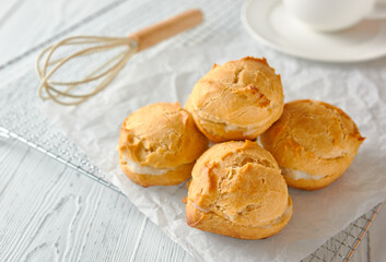 Homemade profiteroles or eclairs on a metal grid. There is a whisk next to it, there is a cup on a saucer behind it. Traditional French dessert. White wooden background. Close-up, copy space.