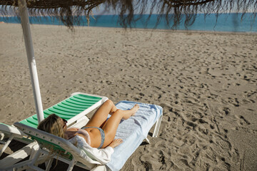 Fototapeta na wymiar Blissful lady in swimwear lying on a deck chair while sunbathing on sandy beach
