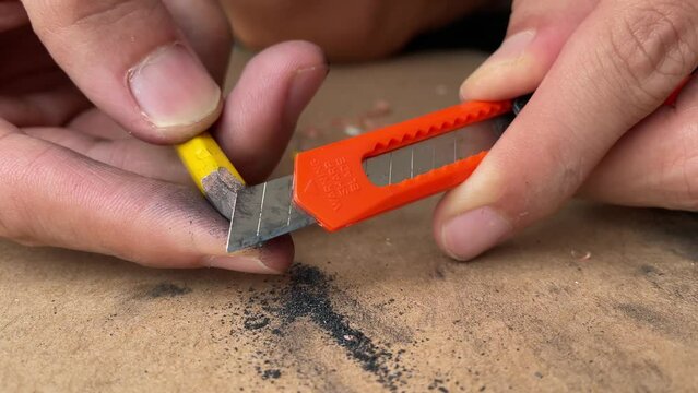 Male hand artist sharpening pencil with a sharp blade box cutter knife close up shot, educational and carpentry concept with cardboard box background - day. Utility razor tool multi purpose uses
