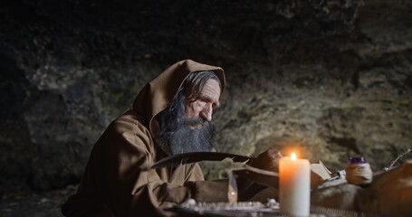 Senior monk in hood sitting in dark rocky cave with burning candle and taking notes with quill