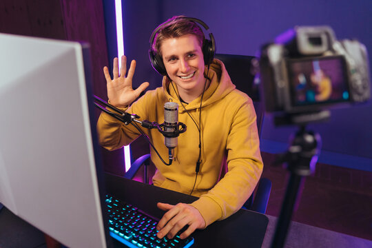 Young handsome vlogger professional gamer waving hand to camera, smiling, greeting his followers with hand gesture while shooting vlog and playing video games on his computer at home. Cyber sport
