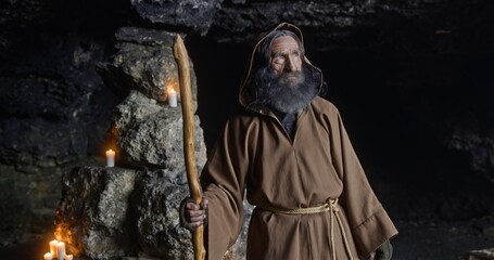 Senior sage man turning around in cave. Elderly bearded male hermit in hooded brown robe with staff turning around and looking at camera near stone walls with candles in cave