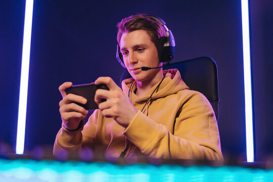 Portrait of caucasian man pro gamer sitting on gaming chair, wearing headset, has live stream while playing mobile game on the smartphone at home. Young cyber sportsman holding cellphone in hands
