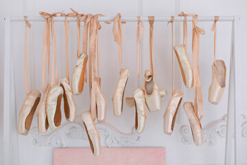 Many hanging ballet shoes on white wall background in studio. New pointe shoes with satin ribbons hanging on rank. Ballet shoes hang on bar in room. Concept of dance, ballet school, ballerinas clothes