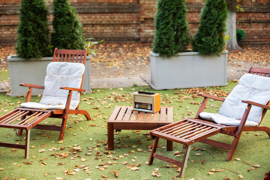 Wooden chairs in autumn garden. Vintage radio on table. Two deckchairs on green summer lawn on picnic. Lounge sunbed. Wooden garden furniture on grass lawn outdoor for relax. Backyard exterior.	
