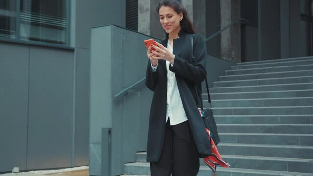 Beautiful young woman caucasian business girl leaving the office using smartphone reading happy timeline social media walking outdoors. Cheerful people. Urban lifestyle.