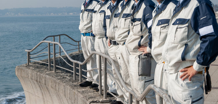 海に向かって腰に手をあてて立つ作業服姿の７人