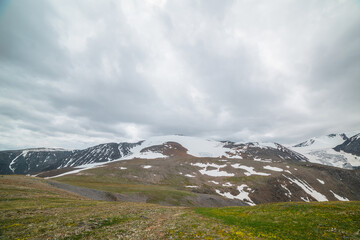 Fototapeta premium Dramatic alpine landscape with large snowy mountain dome in sunlight under gray cloudy sky. Scenic panoramic view to high snow mountain in dome shape in center. Changeable weather at high altitude.