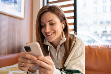 Closeup portrait of a beautiful woman with smiley face holding , using and looking at smartphone and listening music on earphones in modern cafe.
