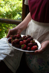 Woman standing at the window and holding ripe strawberry on silver oval plate, summer garden concept, selective focus