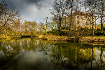 View of a pond in the background of leafless trees and a building reflecting on the water. - obrazy, fototapety, plakaty