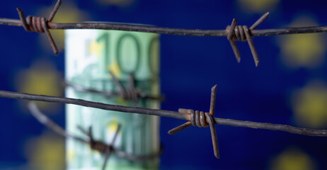 European Union currency wrapped in barbed wire against flag of EU as symbol of Economic warfare, sanctions and embargo busting. Selective focus on barbed wire - obrazy, fototapety, plakaty