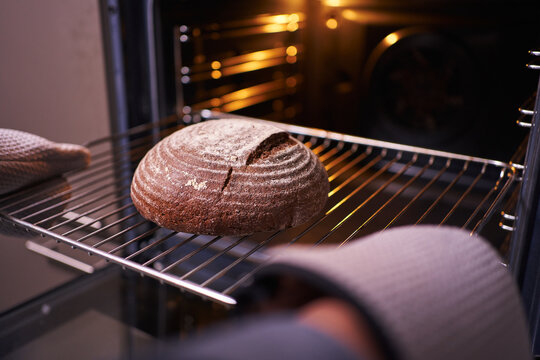 Female Hands In Oven Mitts Take Round Rye Bread Out Of The Oven