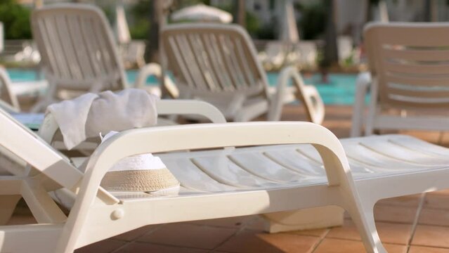 Sun shines on empty white plastic sunbeds at holiday resort, closeup detail, blurred people in near pool behind