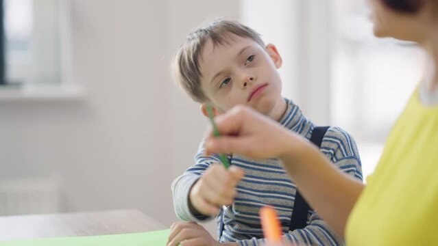 Side view positive autistic boy choosing colors painting in school with unrecognizable tutor. Portrait of Caucasian child with genetic mutation learning art with teacher indoors. Education and autism